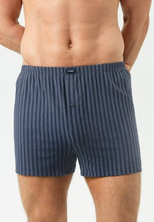 Mann trägt marineblaue gestreifte Boxershorts mit Knopfleiste, steht mit den Händen hinter dem Rücken vor einem einfarbigen hellen Hintergrund.