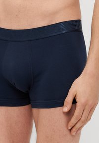 Sininen puuvillasekoitekangas, jossa on teksturoitu joustava vyötärönauha. Ominaisuuksiin kuuluu tiukka istuvuus, sileä pinta ja kontrastivärinen ompelu reunoilla.