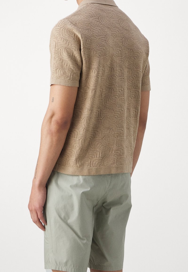 Polo shirt beige a maniche corte in maglia con un motivo a onde texturizzato, abbinato a pantaloni corti verde chiaro, visto da dietro.