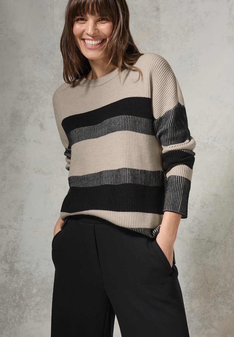 Maglione a righe beige e nere con texture a coste; maniche lunghe e vestibilità rilassata; abbinato a pantaloni neri; indossato da una donna sorridente.