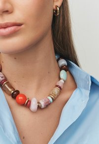 Bunte Perlenkette mit verschiedenen Formen und Größen, ausgestattet mit roten, blauen, pinken und braunen Perlen sowie goldenen Akzenten. Getragen über einem blauen Shirt.