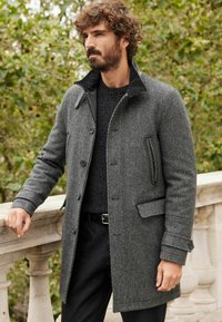 Grå tweedfrakke med en sort krave, knaplukning og sidelommer. Båret over en struktureret sort sweater og sorte bukser.