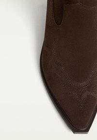 Bottine en daim marron avec un bout pointu, présentant des motifs de couture décoratifs et sans matériel visible. Texture lisse.
