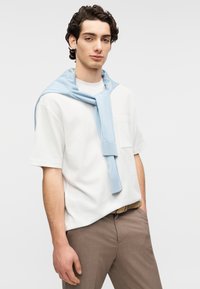 Λευκό κοντομάνικο μπλουζάκι με τσέπη, συνδυασμένο με καφέ παντελόνια και γαλάζιο πουλόβερ δεμένο γύρω από τους ώμους. Λείο ύφασμα, casual σχεδιασμός.