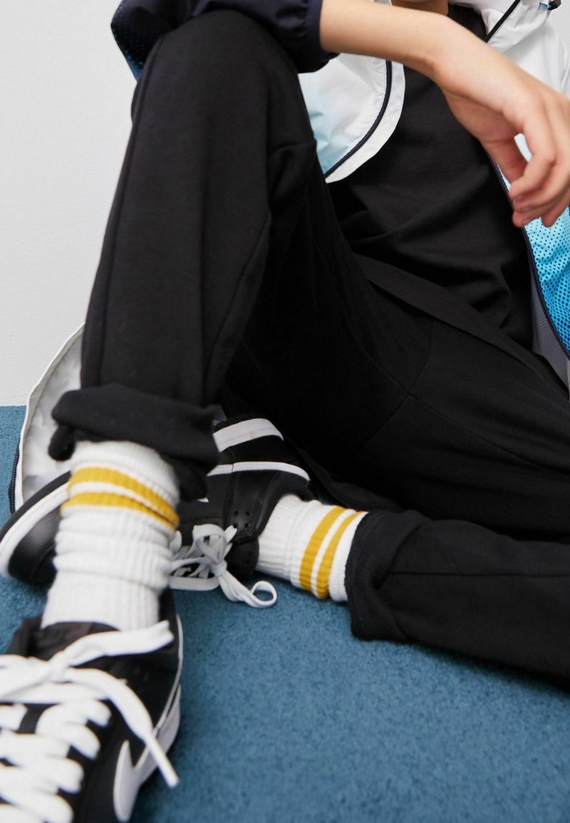 Pantaloni sportivi neri con risvolti, abbinati a calze bianche con strisce gialle e sneakers nere con lacci bianchi. Sfondo blu.