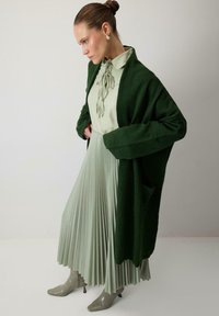 Green knitted cardigan over a light green pleated dress, complemented by glossy gray ankle boots with a pointed toe.