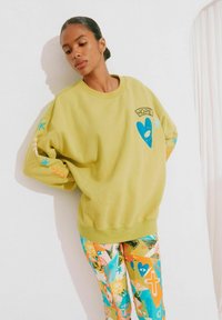 Olivengroene sweatshirt met een blauw hartontwerp met een oog en het woord "HOOP." Gekleed met kleurrijke, motief leggings.