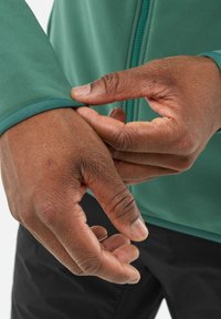Green, lightweight jacket with a textured exterior. Close-up of hands adjusting the cuff, highlighting the smooth fabric and ergonomic fit.