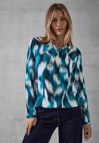 Maglietta a maniche lunghe in un motivo astratto teal e bianco, realizzata in un tessuto morbido, con scollo a V e una vestibilità comoda, abbinata a jeans scuri.