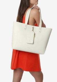 Sac fourre-tout blanc avec de longues poignées, texture lisse, clous décoratifs et une étiquette amovible portant le nom de la marque.