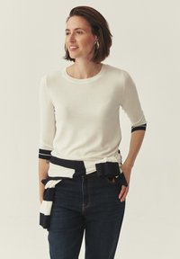 Femme portant une chemise blanche à manches longues et un jean foncé souriant, avec un pull rayé noir et blanc noué autour de la taille, sur un fond uni.
