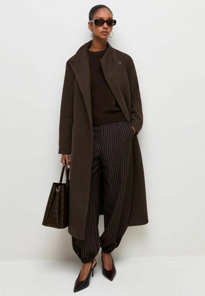 Mujer con abrigo largo marrón oscuro, suéter negro, pantalones de rayas finas, tacones altos negros, gafas de sol, pendientes de aro, llevando bolso oscuro.
