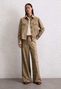 Veste à carreaux marron clair avec poches avant, associée à un pantalon large assorti. L'ensemble est complété par un pull en laine crème et une ceinture marron.