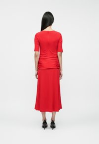 Κόκκινο φόρ εμα μέχρι το γόνατο με εφαρμοστό μπούστο, κοντά μανίκια και ρέουσα φούστα. Διατίθεται με συγκεντρωμένες λεπτομέρειες στο πίσω μέρος. Φοράει μαύρες γόβες.