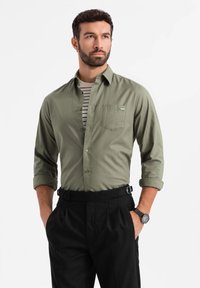 Camicia button-up in verde oliva, in misto cotone, con maniche arrotolate e una tasca sul petto. Indossata sopra una t-shirt a righe con collo a giro, abbinata a pantaloni neri.