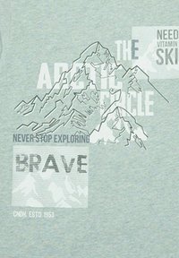 Camiseta de algodón azul claro con un gráfico de montaña, con el texto "EL CÍRCULO ÁRTICO", "NUNCA DEJES DE EXPLORAR" y "VALIENTE" en diferentes fuentes.