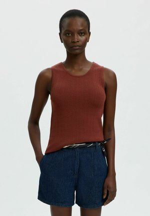 Femme debout face à l'avant, portant un débardeur en maille couleur rouille et un short bleu, une main dans la poche, sur un fond uni clair.