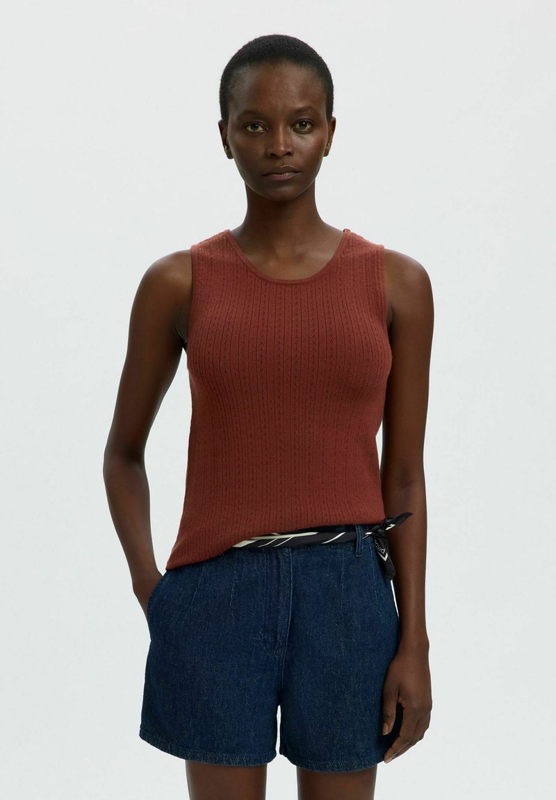 Femme debout face à l'avant, portant un débardeur en maille couleur rouille et un short bleu, une main dans la poche, sur un fond uni clair.