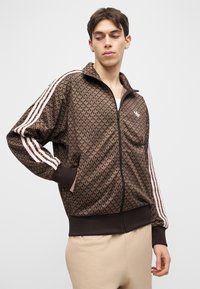 Νέος άνδρας με κοντά σκούρα μαλλιά, φορώντας καφέ ζακέτα με φερμουάρ και ροζ ρίγες στα μανίκια και μπεζ παντελόνι, στέκεται μπροστά σε απλό φόντο.