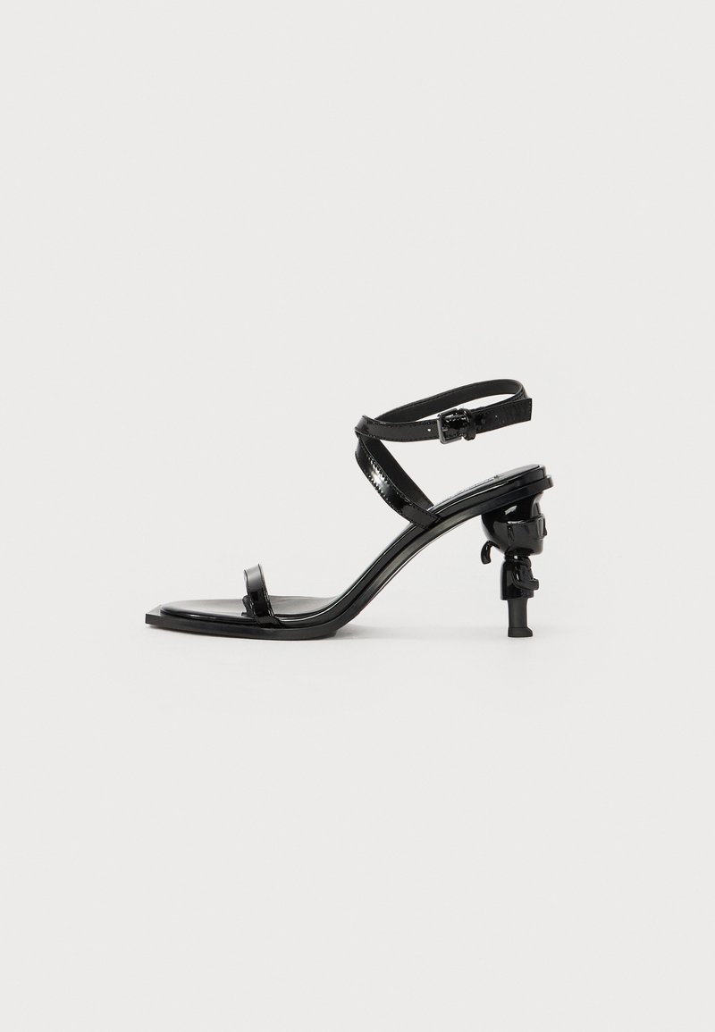 Sandalo con tacco alto in pelle verniciata nera, dotato di due cinturini incrociati e un tacco sottile. Presenta un design del tacco unico e scolpito.