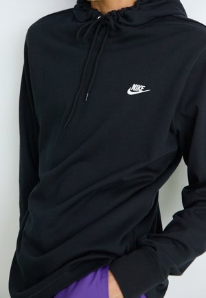 Personne portant un sweat à capuche noir Nike avec cordons et logo blanc, mains partiellement dans les poches d’un pantalon violet.