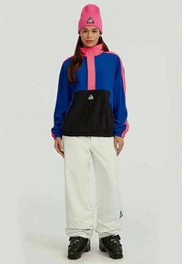 Vrouw in een kleurblok-jack in blauw, roze en zwart, witte broek, roze muts en zwarte winterlaarzen, staande tegen een effen achtergrond.