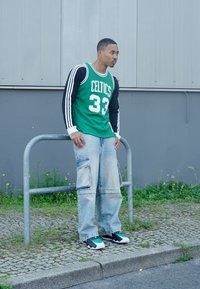 Grünes Basketballtrikot mit weißer Schrift über schwarzen langen Ärmeln, kombiniert mit einer hellen Denim-Cargohose und schwarzen Sneakers mit türkisfarbenen Akzenten.