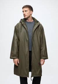 Olīvu zaļš ūdensizturīgs parka ar kapuci, ar teksturētu iekšējo oderējumu, garām piedurknēm un velcro akcentiem uz aprocēm.