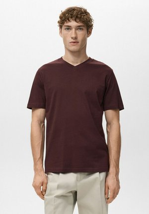 Jonge man met krullend haar, gekleed in een effen donkerbordeauxrood V-hals T-shirt en lichtbeige broek, staand tegen een witte achtergrond.