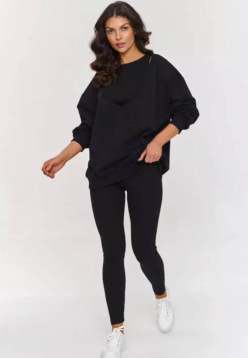 Czarny oversized sweter z opadającymi ramionami, w połączeniu z czarnymi prążkowanymi legginsami. Model nosi białe sneakersy, tworząc monochromatyczny wygląd.
