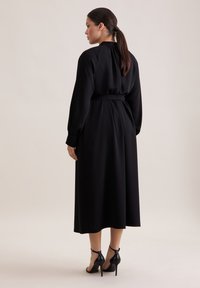 Schwarz-midi Kleid mit langen Ärmeln, einem Bindegürtel und einer glatten Textur. Getragen mit schwarzen High-Heels. Rückansicht gezeigt.