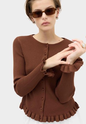 Person trägt braunen, gerippten, geknöpften Cardigan mit Rüschen an Bündchen und Saum, braune rechteckige Sonnenbrille, goldene Creolen und einen Ring.