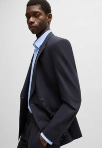 Blazer navy, design doppiopetto, revers a punte, bottoni neri, indossato su una camicia a quadri azzurra chiara, tessuto liscio, vestibilità sagomata.