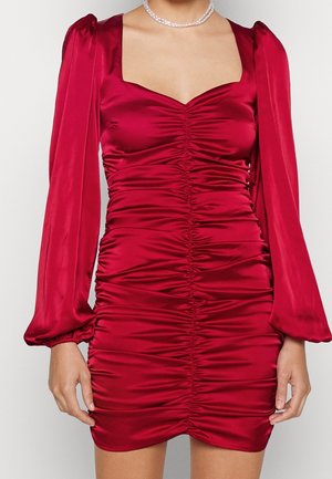 Femme portant une robe rouge en satin cintrée et froncée, avec des manches longues bouffantes et un décolleté carré, accessoirisée d'un collier de perles.