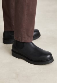 Bottines en cuir noir avec panneaux élastiques sur les côtés, bout rond et semelle en caoutchouc texturé, associées à un chino marron.