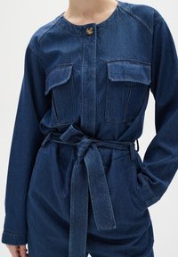 Denim overall v tmavě modré barvě s knoflíkovým zapínáním, dvěma náprsními kapsami a páskem vázaným v pase. Obsahuje kulatý výstřih a dlouhé rukávy.