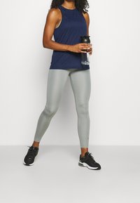Marinblå tank top med en texturerad design, tillsammans med ljusgrå leggings. Modellen håller en klar vattenflaska. Svarta träningsskor fulländar outfiten.