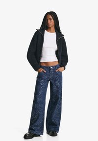 Mornarsko pleteno zip-jakna; bijela pripijena majica; široke hlače s plavim štrasom. Model stoji s rukama u džepovima, crne cipele.