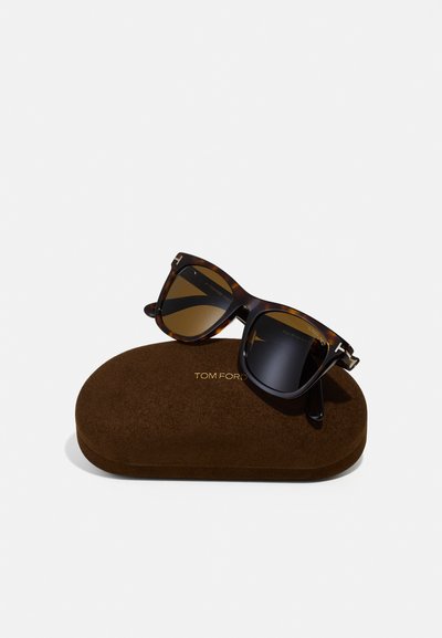 Ochelari de soare cu model țestoasă, cu lentile degradate, așezati pe un toc maro, cu atingere delicată. Logo-ul auriu "TOM FORD" pe toc.