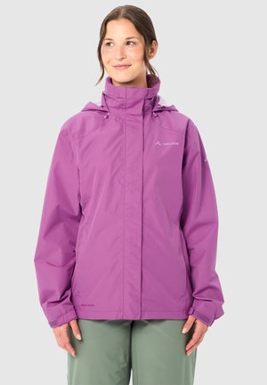 Frau trägt eine violette wasserdichte Jacke mit hohem Kragen und Kapuze, grüne Hose, steht vor einem einfarbigen hellgrauen Hintergrund.