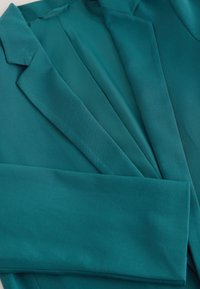 Veste blazer bleu sarcelle pliée à plat montrant les revers, le col et une partie d'une manche.