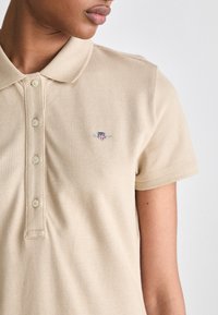Beige polo skjorte laget av strukturert stoff, med krage, fem knapper og et broderi på venstre bryst. Korte ermer.