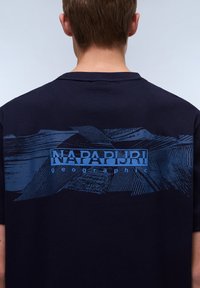 T-shirt bleu marine avec un grand graphisme bleu au dos, mettant en avant le nom de la marque "NAPAPIJRI" et des motifs rayés abstraits. Manches courtes, col rond.