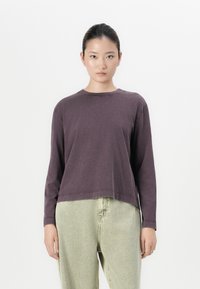 Jonge vrouw met haar opgestoken, gekleed in een donkerpaars shirt met lange mouwen en lichtgroene, ruime jeans met hoge taille, staande voor een witte achtergrond.