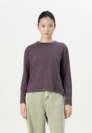 Ung kvinne med hår bunnet opp iført en langermet mørk lilla skjorte og høyhåndte lysegrønne løse jeans, stående mot hvit bakgrunn.