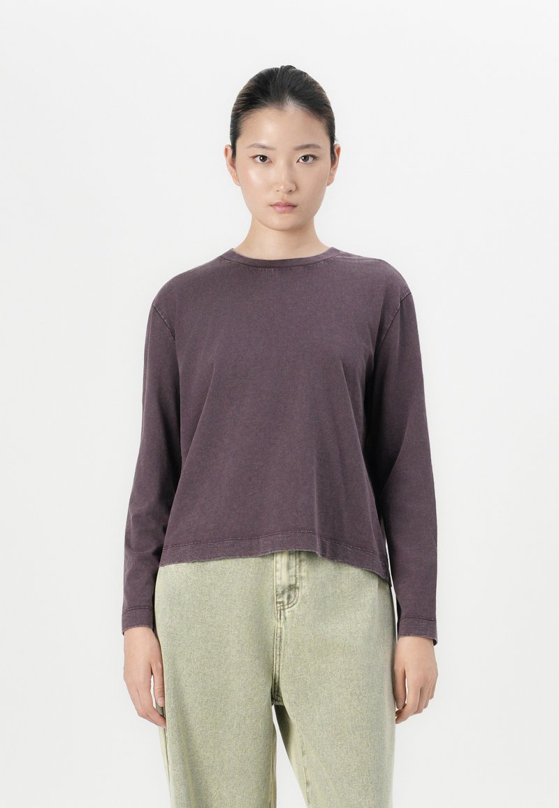 Young woman with hair tied back wearing a long-sleeve dark purple shirt and high-waisted light green loose jeans, standing against white background.