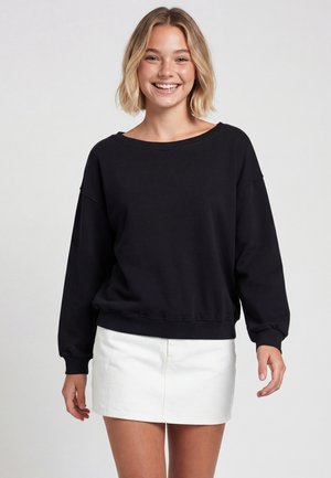 Ung kvinde smiler iført en sort langærmet sweatshirt og en hvid mininederdel, stående mod en ensfarvet baggrund.