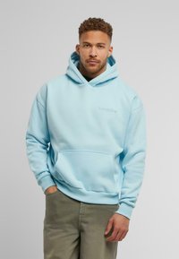 Lichtblauwe hoodie van zacht materiaal, met een voorzak en een capuchon met trekkoord. Subtiele geborduurde branding op de borst.