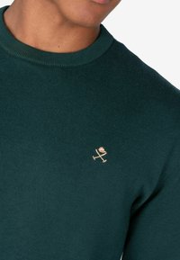 Dunkelgrüner gestrickter Pullover mit Rundhalsausschnitt und einem kleinen gestickten Logo in Hellbraun im linken Brustbereich.