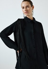 Black button-up shirt with long, textured fringe detailing on sleeves. Made from smooth fabric, featuring a classic collar and straight cut.